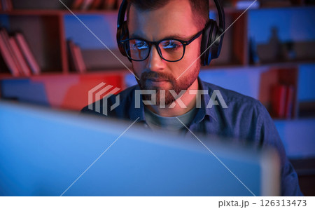 Front view. Man is sitting by a computer indoors at home. Neon lighting 126313473
