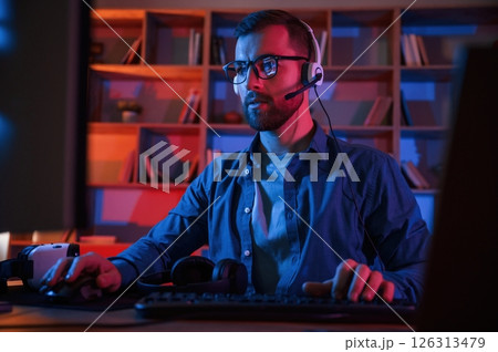 Front view. Man is sitting by a computer indoors at home. Neon lighting Front view. Man is sitting by a computer indoors at home. Neon lighting 126313479