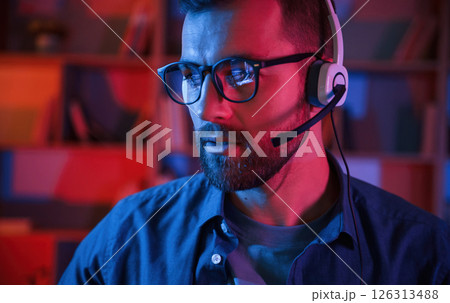 Portrait of man that is sitting by a computer indoors at home. Neon lighting Portrait of man that is sitting by a computer indoors at home. Neon lighting 126313488