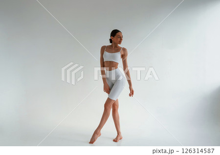 In white fitness clothes. Young caucasian woman with slim body shape is indoors in the studio 126314587