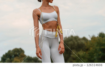 Holding jumping rope. Young caucasian woman with slim body shape is in the fitness clothes outdoors Holding jumping rope. Young caucasian woman with slim body shape is in the fitness clothes outdoors 126314608