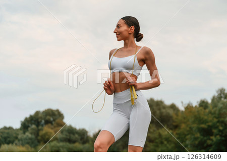 Holding jumping rope. Young caucasian woman with slim body shape is in the fitness clothes outdoors Holding jumping rope. Young caucasian woman with slim body shape is in the fitness clothes outdoors 126314609