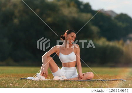 Enjoying nature. Sitting on the mat. Young caucasian woman with slim body shape is in the fitness clothes outdoors 126314643