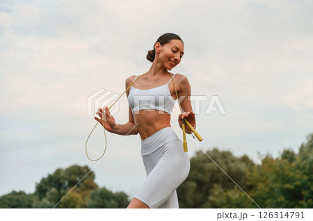 Holding jumping rope. Young caucasian woman with slim body shape is in the fitness clothes outdoors 126314791