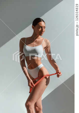 Using the measuring tape. Young caucasian woman with slim body shape is indoors in the studio Using the measuring tape. Young caucasian woman with slim body shape is indoors in the studio 126314819