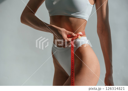 Using measuring tape on legs. Young caucasian woman with slim body shape is indoors in the studio 126314821