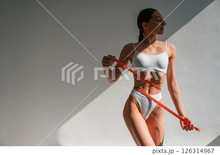 Red colored measuring tape in hands. Young caucasian woman with slim body shape is indoors in the studio 126314967