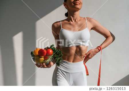Standing against wall and holding healthy food. Young caucasian woman with slim body shape is indoors in the studio Standing against wall and holding healthy food. Young caucasian woman with slim body shape is indoors in the studio 126315013