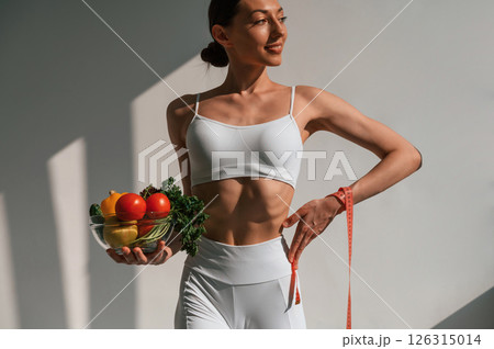 Standing against wall and holding healthy food. Young caucasian woman with slim body shape is indoors in the studio 126315014
