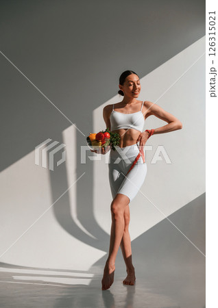 Standing in the sunlight. Holding food. Young caucasian woman with slim body shape is indoors in the studio Standing in the sunlight. Holding food. Young caucasian woman with slim body shape is indoors in the studio 126315021