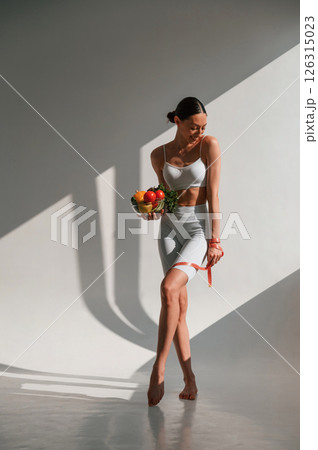 Standing in the sunlight. Holding food. Young caucasian woman with slim body shape is indoors in the studio Standing in the sunlight. Holding food. Young caucasian woman with slim body shape is indoors in the studio 126315023