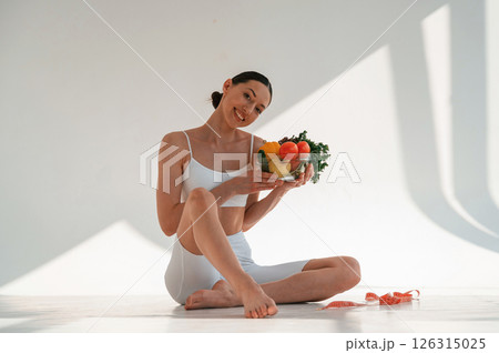Sitting on the floor with healthy food. Young caucasian woman with slim body shape is indoors in the studio Sitting on the floor with healthy food. Young caucasian woman with slim body shape is indoors in the studio 126315025