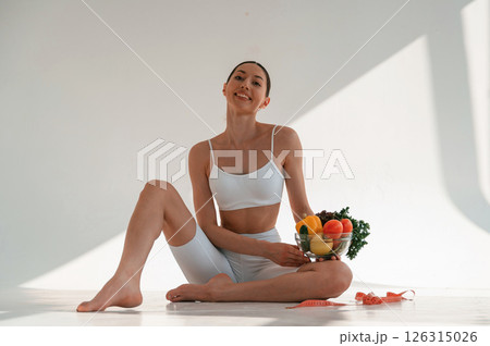 Sitting on the floor with healthy food. Young caucasian woman with slim body shape is indoors in the studio Sitting on the floor with healthy food. Young caucasian woman with slim body shape is indoors in the studio 126315026