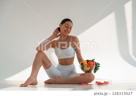 Sitting on the floor with healthy food. Young caucasian woman with slim body shape is indoors in the studio Sitting on the floor with healthy food. Young caucasian woman with slim body shape is indoors in the studio 126315027