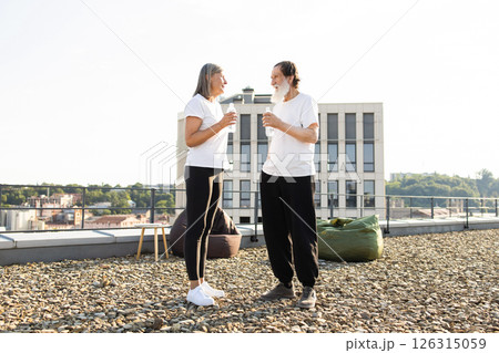 Senior man and woman standing on urban rooftop drinking bottled water, wearing casual clothing outdoors under sunlight Senior man and woman standing on urban rooftop drinking bottled water, wearing casual clothing outdoors under sunlight 126315059