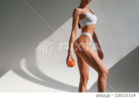 With an orange in hand. Young caucasian woman with slim body shape is indoors in the studio 126315114