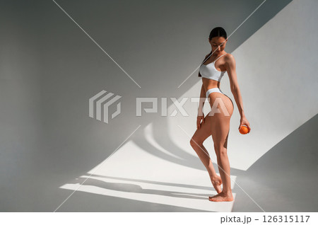 Young caucasian woman with slim body shape is indoors in the studio. Holding an orange 126315117