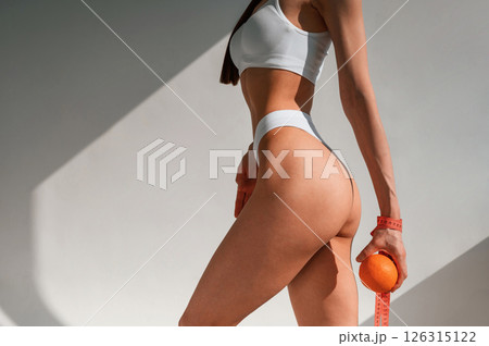 Standing, posing and holding an orange in hand. Young caucasian woman with slim body shape is indoors in the studio Standing, posing and holding an orange in hand. Young caucasian woman with slim body shape is indoors in the studio 126315122