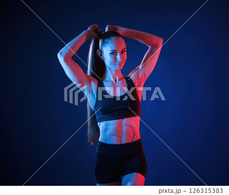 Fixes her hair. Beautiful muscular woman is indoors in the studio with neon lighting Fixes her hair. Beautiful muscular woman is indoors in the studio with neon lighting 126315383
