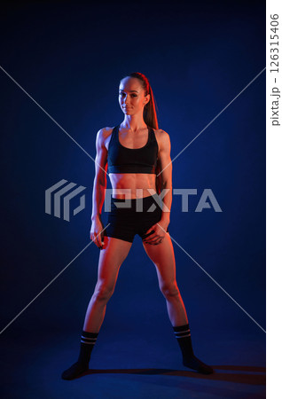 Front view, standing. Beautiful muscular woman is indoors in the studio with neon lighting Front view, standing. Beautiful muscular woman is indoors in the studio with neon lighting 126315406
