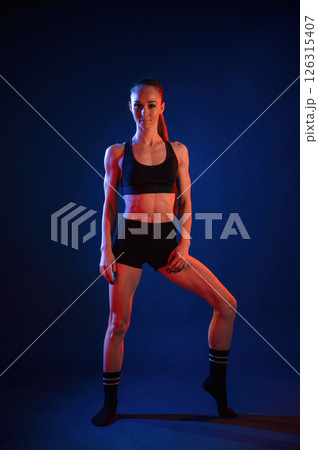 Front view, standing. Beautiful muscular woman is indoors in the studio with neon lighting Front view, standing. Beautiful muscular woman is indoors in the studio with neon lighting 126315407
