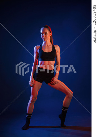 Front view, standing. Beautiful muscular woman is indoors in the studio with neon lighting Front view, standing. Beautiful muscular woman is indoors in the studio with neon lighting 126315408
