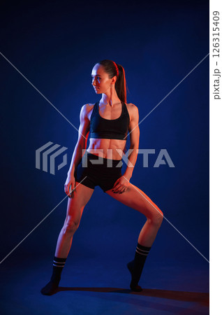 Front view, standing. Beautiful muscular woman is indoors in the studio with neon lighting Front view, standing. Beautiful muscular woman is indoors in the studio with neon lighting 126315409