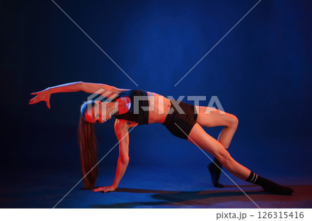 Exercise by standing on a leg and hand. Beautiful muscular woman is indoors in the studio with neon lighting Exercise by standing on a leg and hand. Beautiful muscular woman is indoors in the studio with neon lighting 126315416