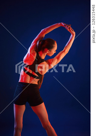 Flexible body. Beautiful muscular woman is indoors in the studio with neon lighting Flexible body. Beautiful muscular woman is indoors in the studio with neon lighting 126315448