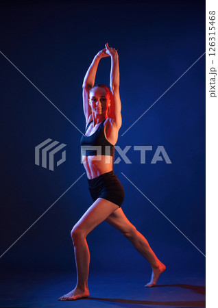 Sportive exercises, standing and stretching the hands. Beautiful muscular woman is indoors in the studio with neon lighting 126315468