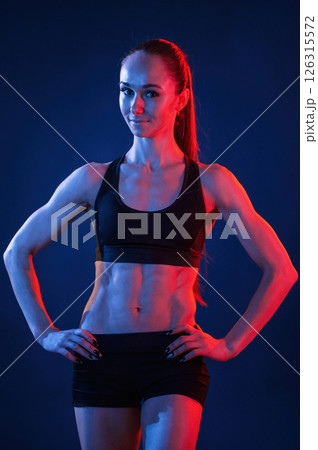 Standing with the hands on waist. Beautiful muscular woman is indoors in the studio with neon lighting 126315572