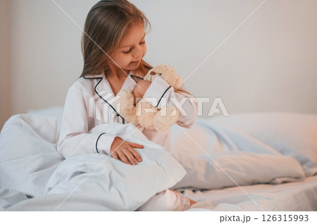 Little girl with teddy bear in hands is sitting on the bed at home Little girl with teddy bear in hands is sitting on the bed at home 126315993