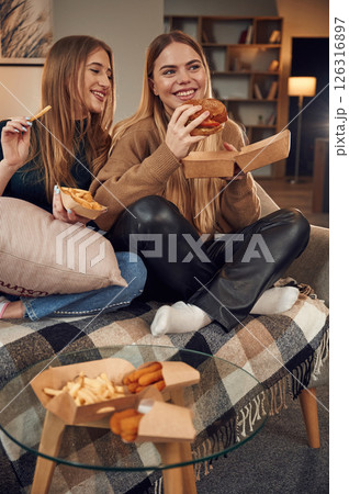 Smiling, enjoying the meal. Two women are sitting and eating burger, French fries and chicken nuggets indoors together 126316897