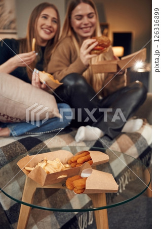 Smiling, enjoying the meal. Two women are sitting and eating burger, French fries and chicken nuggets indoors together 126316899
