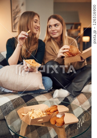 Two women are sitting and eating burger, French fries and chicken nuggets indoors together 126316900