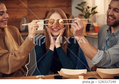 Having fun, rolls of the food in the front of the eyes of woman. Friends are sitting indoors at home and eating sushi together Having fun, rolls of the food in the front of the eyes of woman. Friends are sitting indoors at home and eating sushi together 126317140