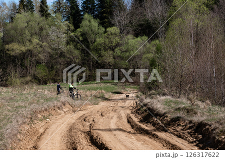 Two cyclists pause by a dirt road, surrounded by lush greenery and a tranquil forest. 126317622