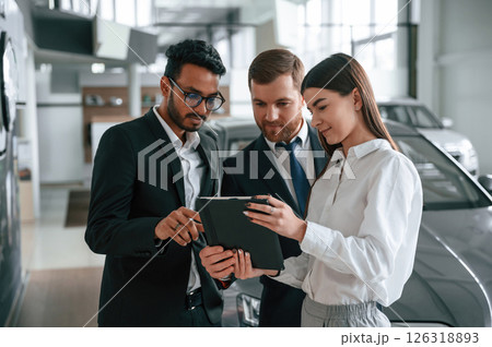 Standing and reading information in the graphic tablet. Three people are working together in the car showroom 126318893