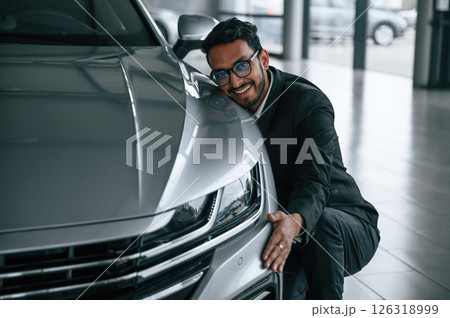 Embracing the transport. Handsome indian man in suit is in the car showroom with the vehicle indoors 126318999