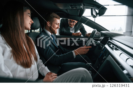 Side view. Car showroom worker is consulting man and woman that are sitting inside of automobile 126319431