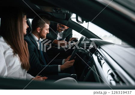 Car showroom worker is consulting man and woman that are sitting inside of automobile Car showroom worker is consulting man and woman that are sitting inside of automobile 126319437