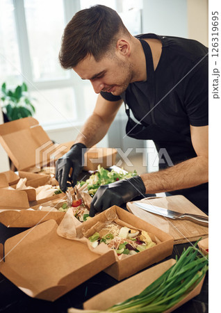 Vegetables added to the burgers. Man is packing food into the paper eco boxes. Indoors, restaurant Vegetables added to the burgers. Man is packing food into the paper eco boxes. Indoors, restaurant 126319695