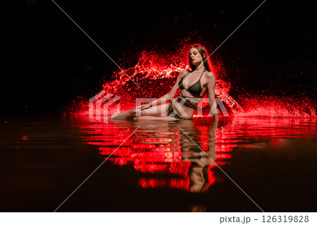 Young slender girl in a swimsuit posing in the water at sunset at the background of red colored water droplets flying in the air 126319828