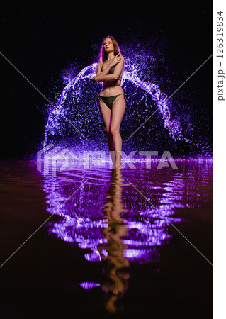 Young slender girl in a swimsuit posing in the water at sunset at the background of purple colored water droplets flying in the air 126319834