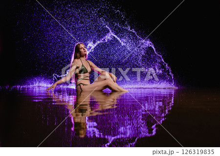 Young slender girl in a swimsuit posing in the water at sunset at the background of purple colored water droplets flying in the air 126319835