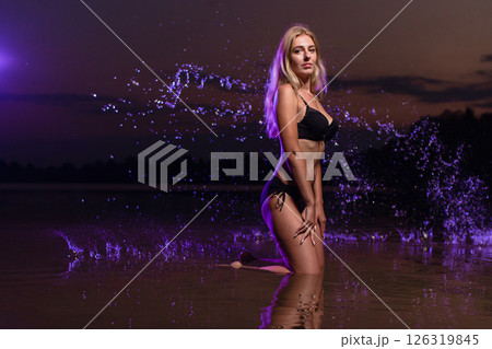 Young slender girl in a swimsuit posing in the water at sunset at the background of purple colored water droplets flying in the air 126319845