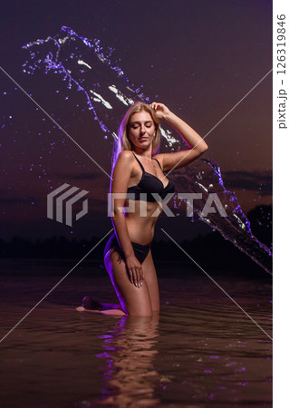 Young slender girl in a swimsuit posing in the water at sunset at the background of purple colored water droplets flying in the air Young slender girl in a swimsuit posing in the water at sunset at the background of purple colored water droplets flying in the air 126319846