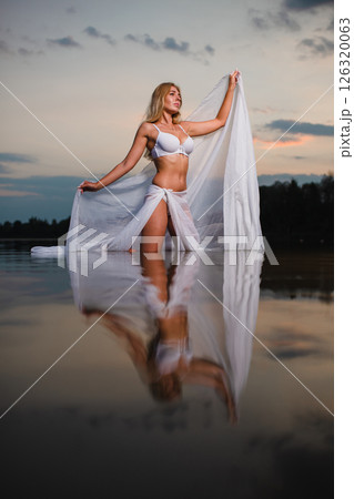 Young slender girl in a swimsuit posing in the water at sunset with big piece of fabric 126320063