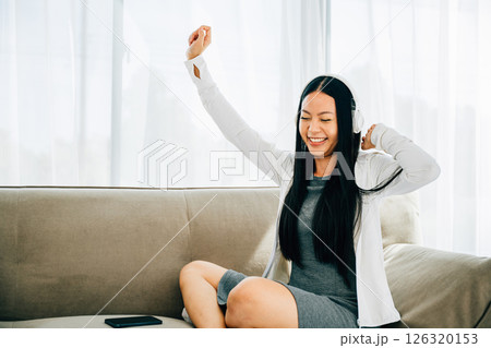 Seated comfortably on the sofa a woman holds her smartphone dancing with headphones on. Immersed in music experiencing happiness and leisurely entertainment at home. Lively relaxation concept. Seated comfortably on the sofa a woman holds her smartphone dancing with headphones on. Immersed in music experiencing happiness and leisurely entertainment at home. Lively relaxation concept. 126320153