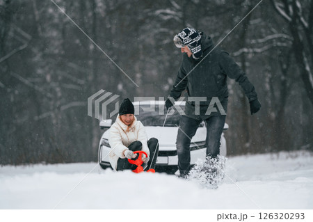 Happy couple are outdoors at winter time. Man takes his wife on a sled. White car behind 126320293